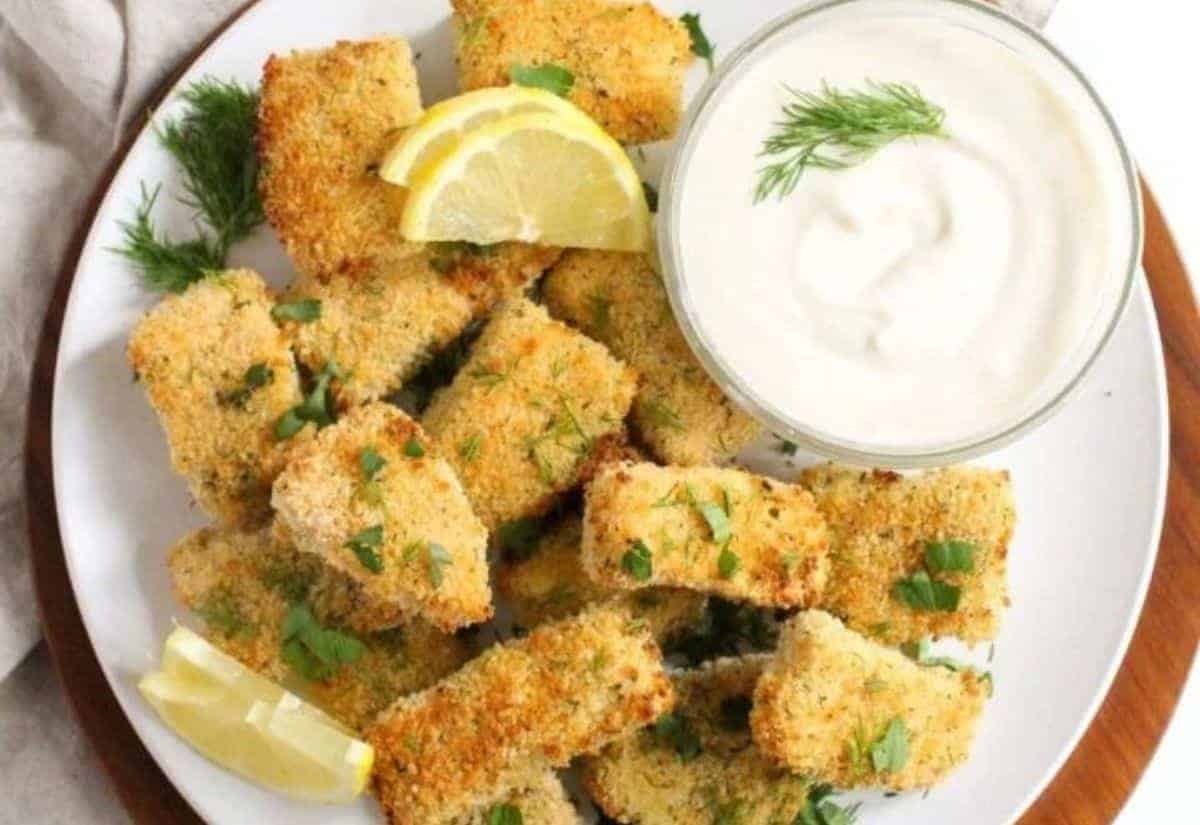 A plate of breaded fish nuggets garnished with fresh herbs and lemon wedges, accompanied by a bowl of creamy dip topped with dill.
