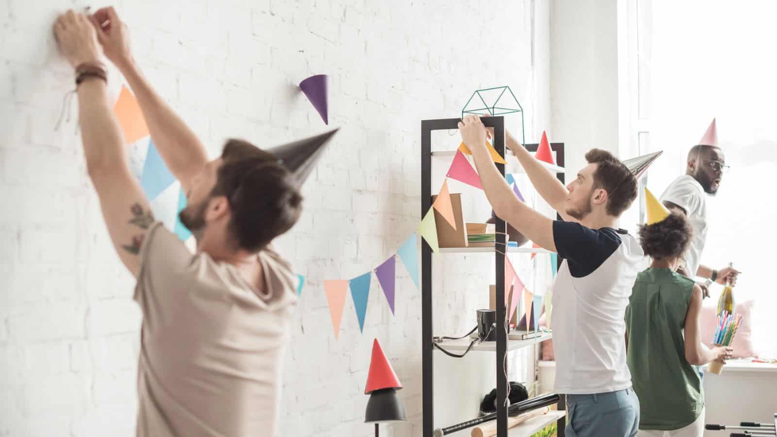 Four people are decorating a white brick wall with colorful paper bunting flags. They are wearing party hats. A bookshelf with small decorations is visible in the background.