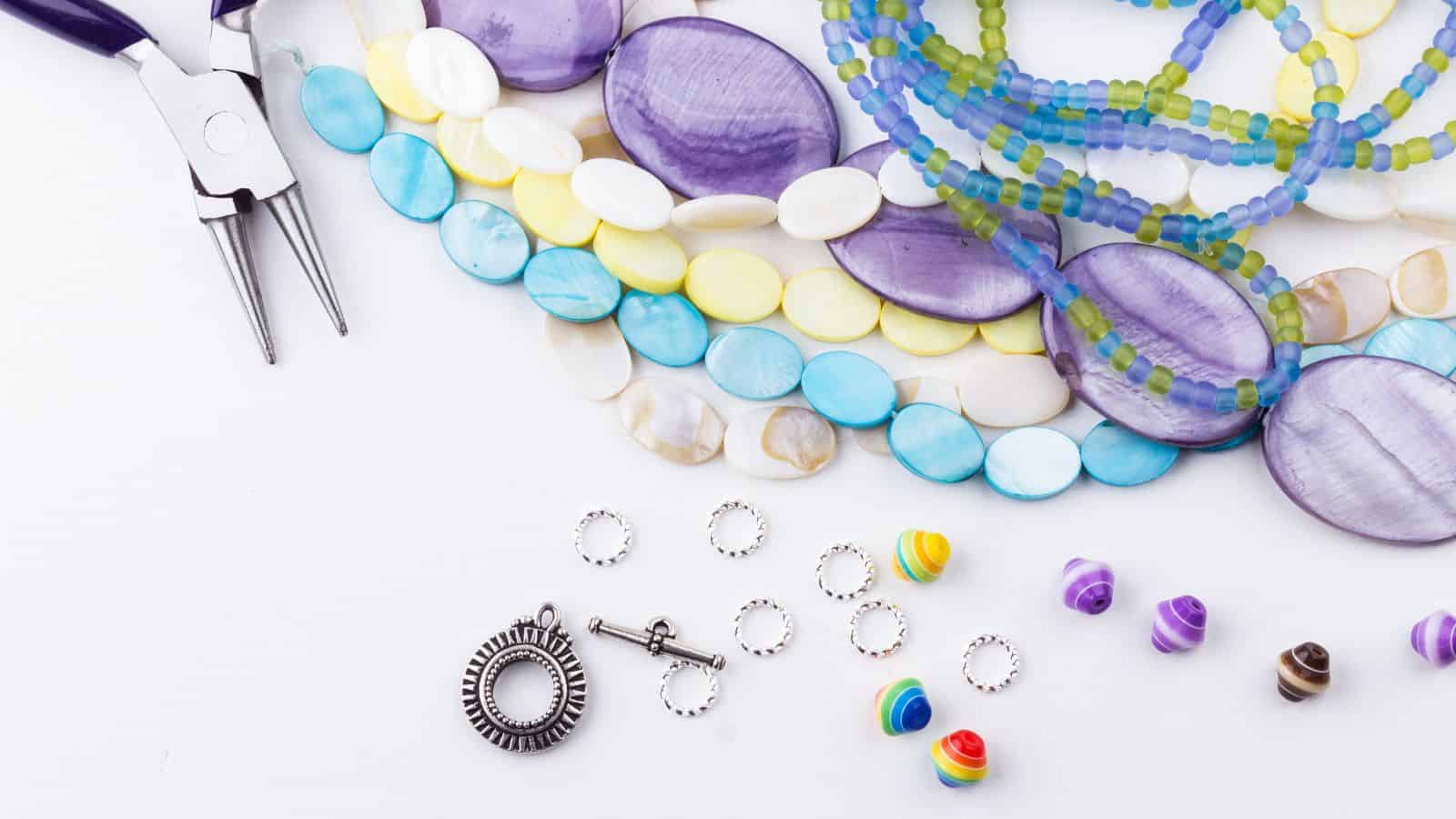 Colorful beads and jewelry-making tools are arranged on a white surface. There are purple, yellow, blue, and multicolored beads along with a pair of pliers, silver toggles, and jump rings.