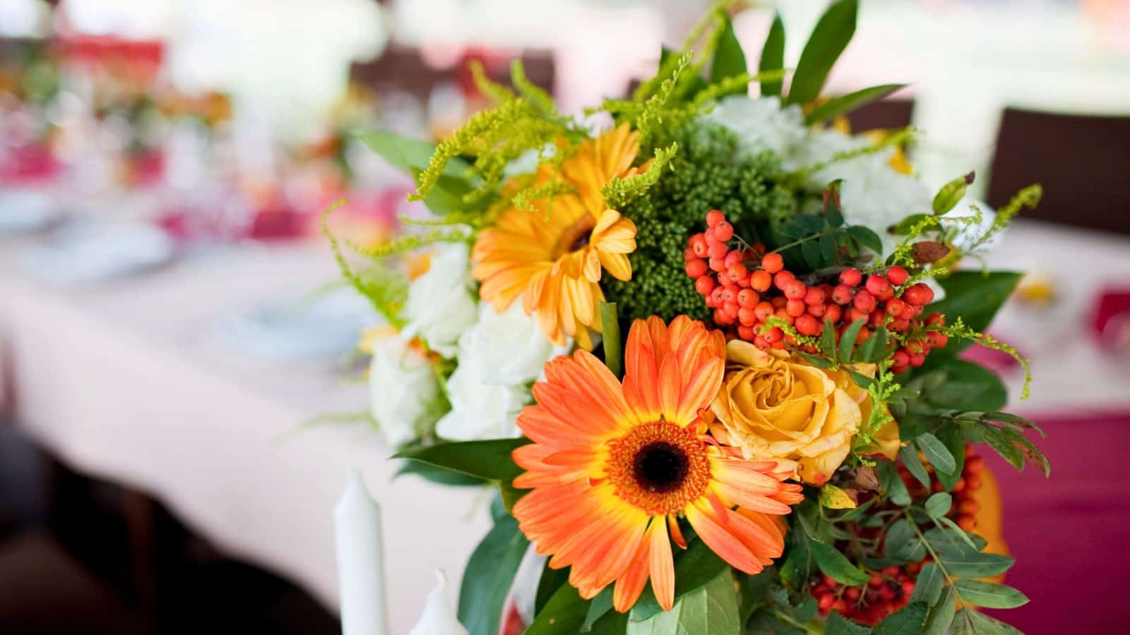A vibrant floral arrangement featuring orange gerbera daisies, yellow roses, red berries, and various green leaves is displayed. The bouquet is set on a table covered with a white and burgundy tablecloth, with blurred table settings in the background.