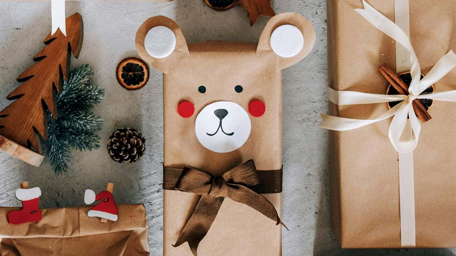 Brown paper gift wrap decorated with bear face, red cheeks, and brown bow. Nearby are a pinecone, a small fir branch, dried orange slices, a wooden tree, and a reindeer decorated gift, creating a festive atmosphere.