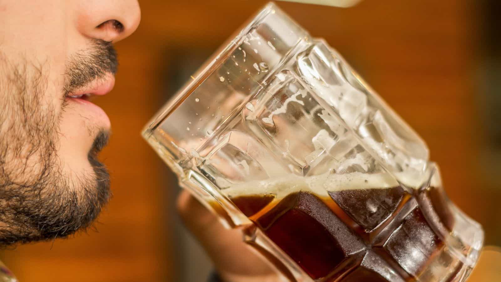 A person with facial hair is drinking a dark beverage from a large, transparent glass mug. The background is out of focus, emphasizing the drink and the action.