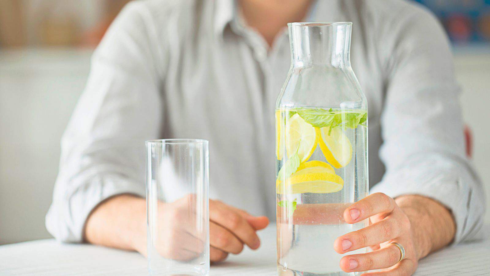 A person wearing a light shirt sits at a table with a clear glass and a carafe filled with water, lemon slices, and mint. The person's hand is holding the carafe. The background is softly blurred.