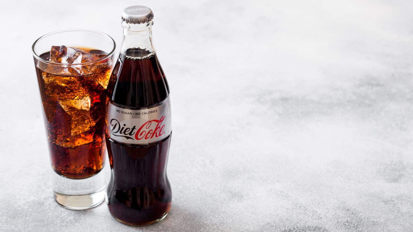 A glass of iced Diet Coke is placed next to a bottle of Diet Coke on a light gray textured surface. The glass is filled with ice cubes and the bottle is capped with a silver top.