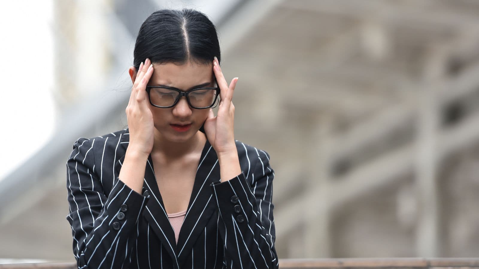 A person wearing glasses and a striped blazer holds their head with both hands, looking downwards. They appear to be in a state of concentration or stress. The background is blurred, possibly indicating an outdoor setting.