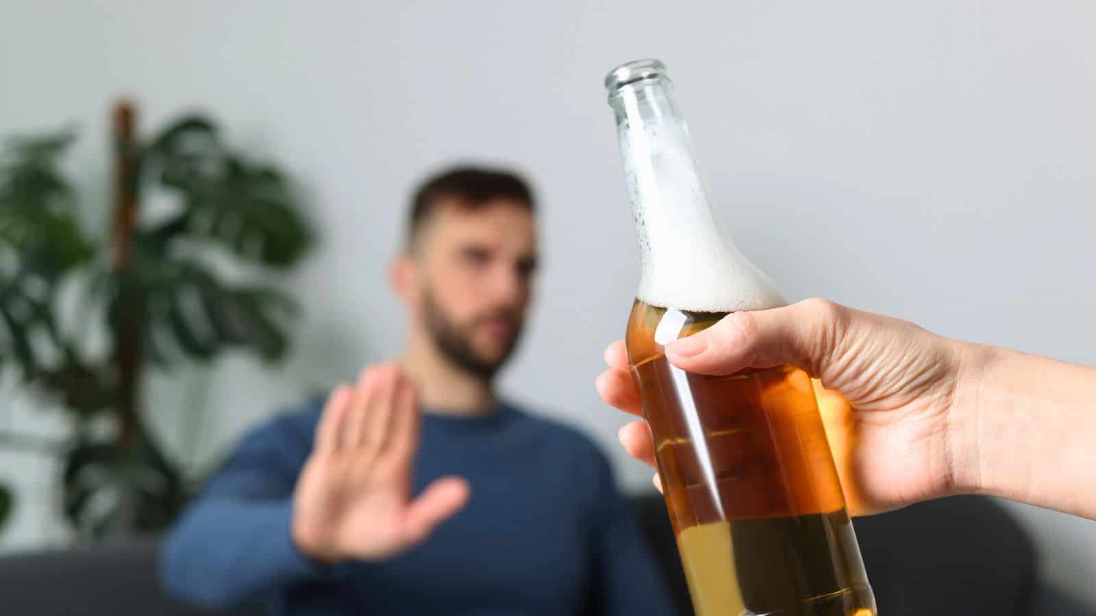A person sitting on a couch extends their hand in a gesture of refusal towards another person holding a beer bottle in the foreground. A large green plant is partially visible in the background.