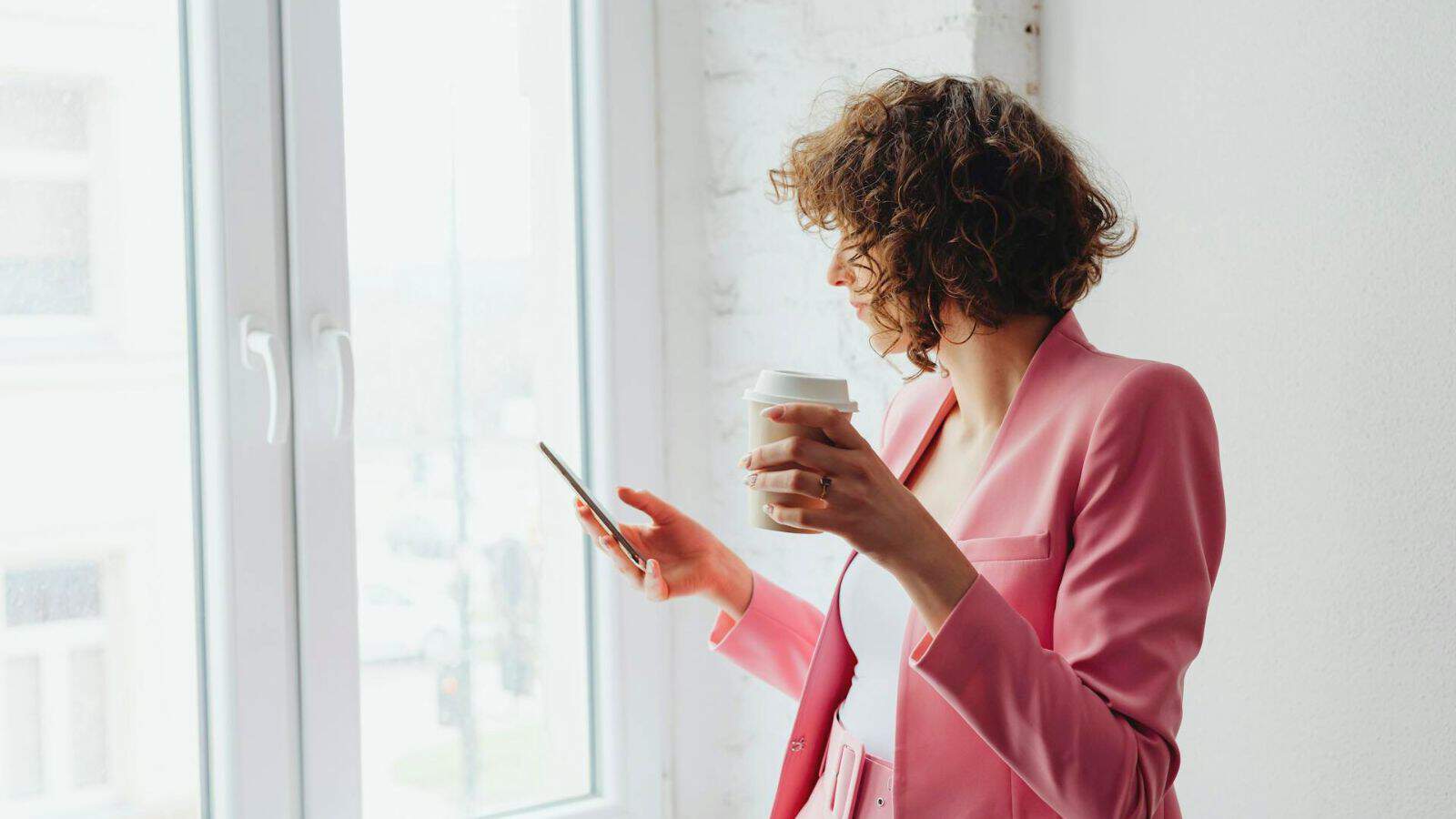 A person with curly hair is standing by a bright window, holding a smartphone in one hand and a disposable coffee cup in the other. They are wearing a pink blazer and looking out of the window.