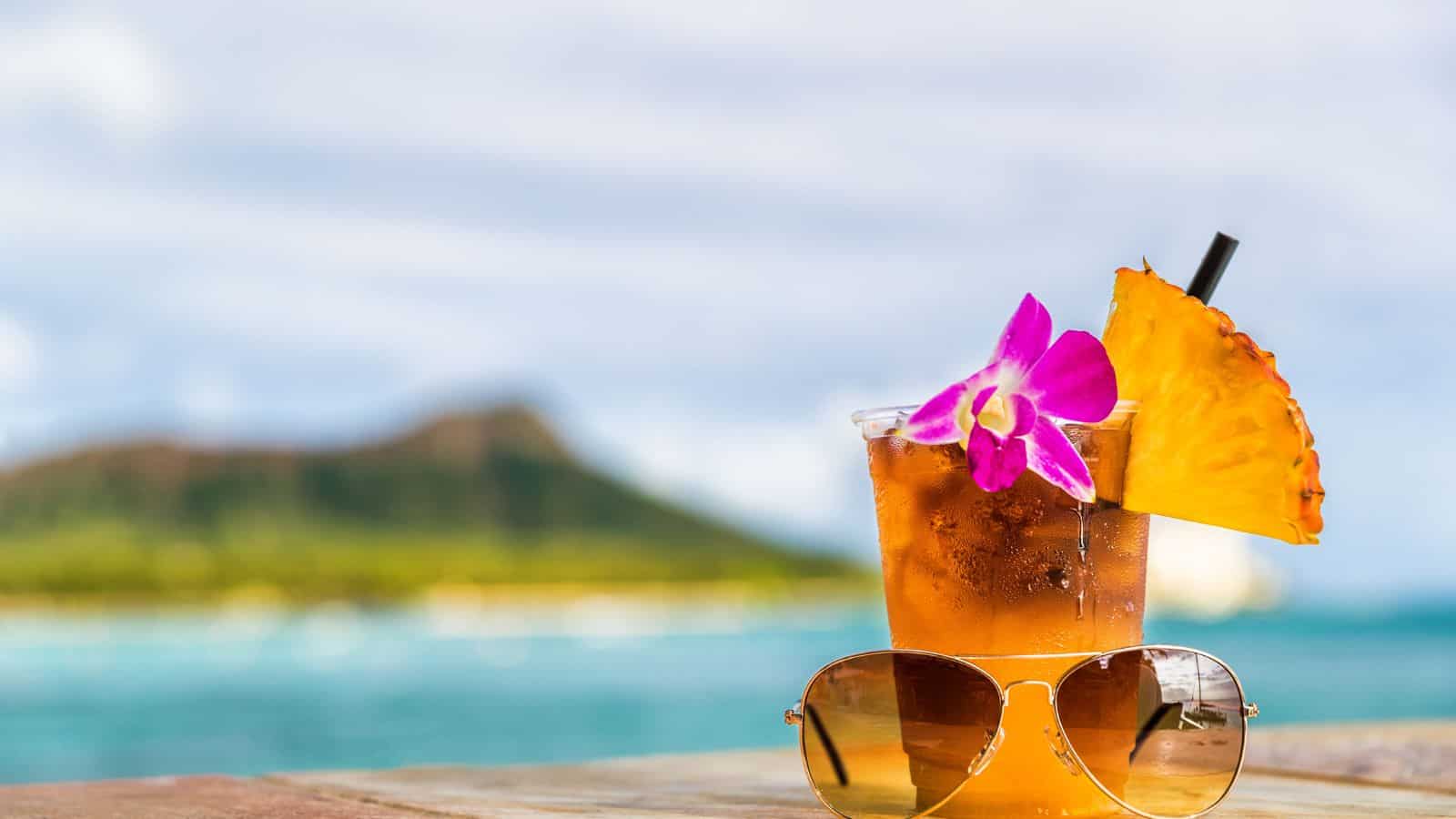 A tropical drink garnished with a purple orchid and a pineapple slice sits on a table. In front of the drink are a pair of sunglasses. The background shows the ocean and a distant, blurred view of a green mountain.