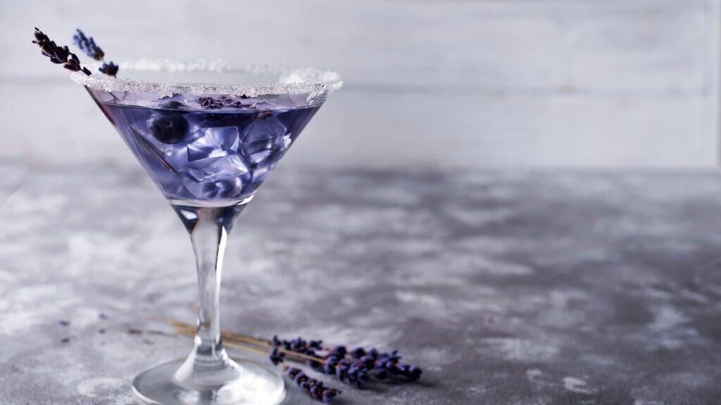 A purple cocktail in a martini glass with sugar on the rim and ice cubes inside. Garnished with a sprig of lavender and two blueberries. Two sprigs of lavender lie next to the glass on a gray textured surface.