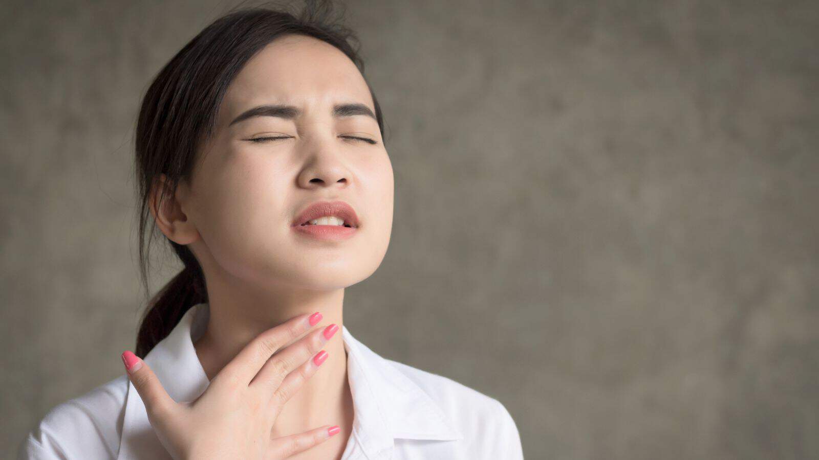 A person with dark hair and a white shirt touches their throat, appearing to be in discomfort. The person's eyes are closed, and they have a pained expression. The background is a plain, muted color.