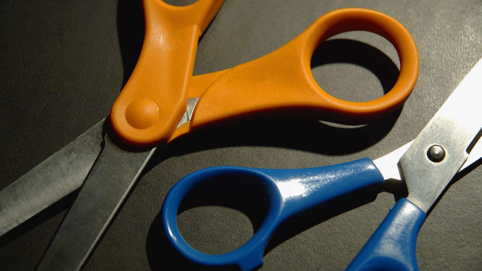 Two pairs of scissors on a dark surface, one with orange handles and the other with blue handles. The scissors are positioned with their blades slightly overlapping.