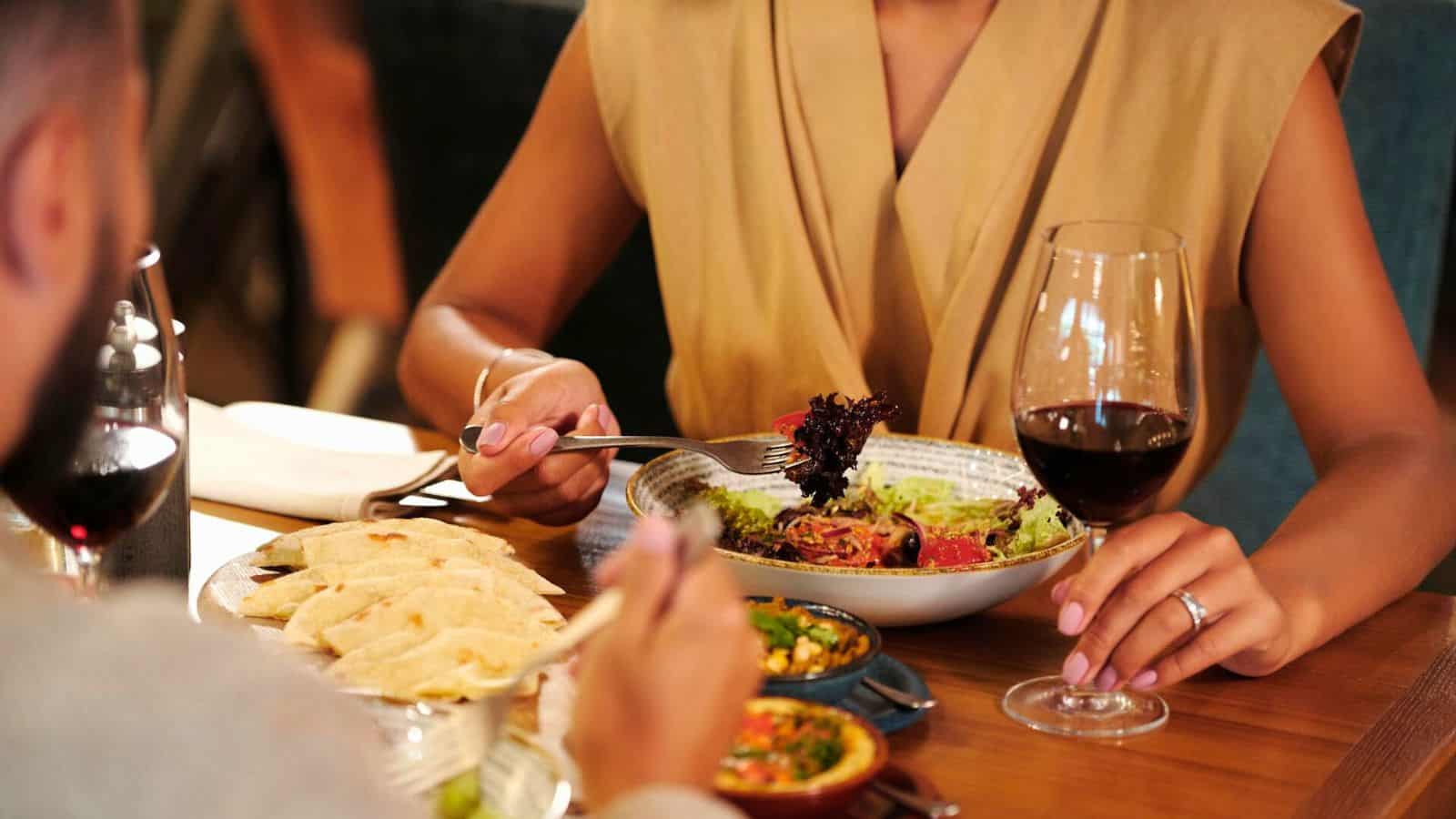 Two people dining at a table. One person is holding a fork with salad from a bowl. There is a glass of red wine nearby. The other person is holding a fork and is about to eat from a plate with bread and dishes. The atmosphere is warm and inviting.