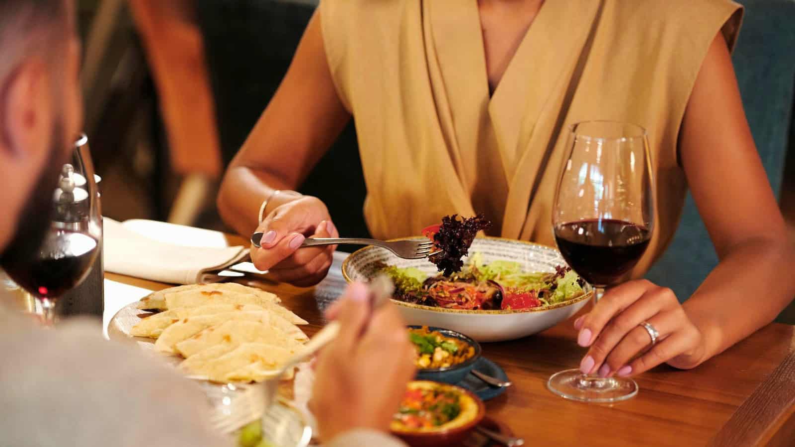 Two people dining at a table with a focus on the food. One person has a plate of salad and a glass of red wine, while the other has a plate of folded tortillas. Small bowls of dips are visible on the table.