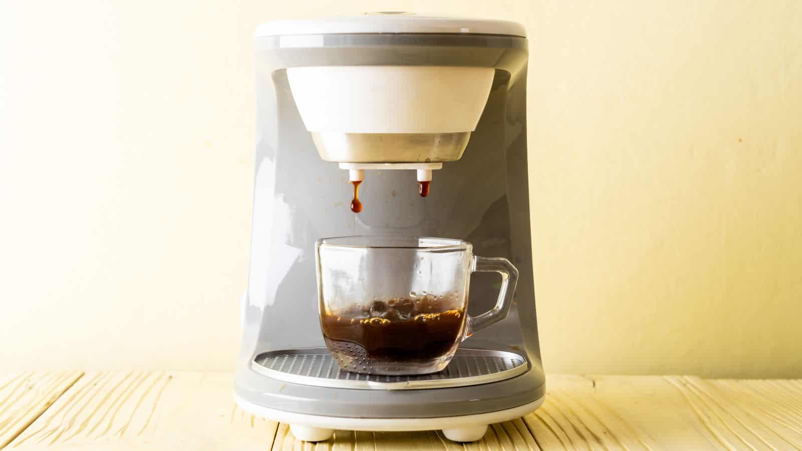 A single-serve coffee machine dispensing coffee into a clear glass mug on a wooden surface. The machine is grey with white accents, and the background is a plain light yellow wall.