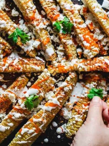 Grilled corn ribs on a dark surface, topped with crumbled cheese, sour cream, and a drizzle of hot sauce. A hand is adding cilantro leaves as a garnish.
