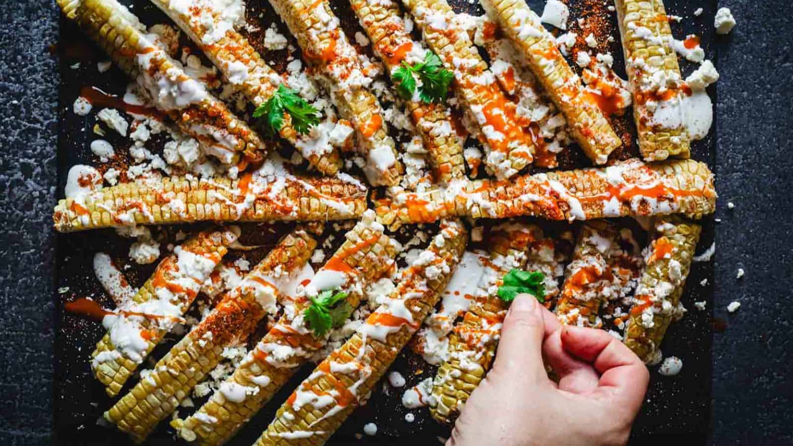 Grilled corn ribs on a dark surface, topped with crumbled cheese, sour cream, and a drizzle of hot sauce. A hand is adding cilantro leaves as a garnish.