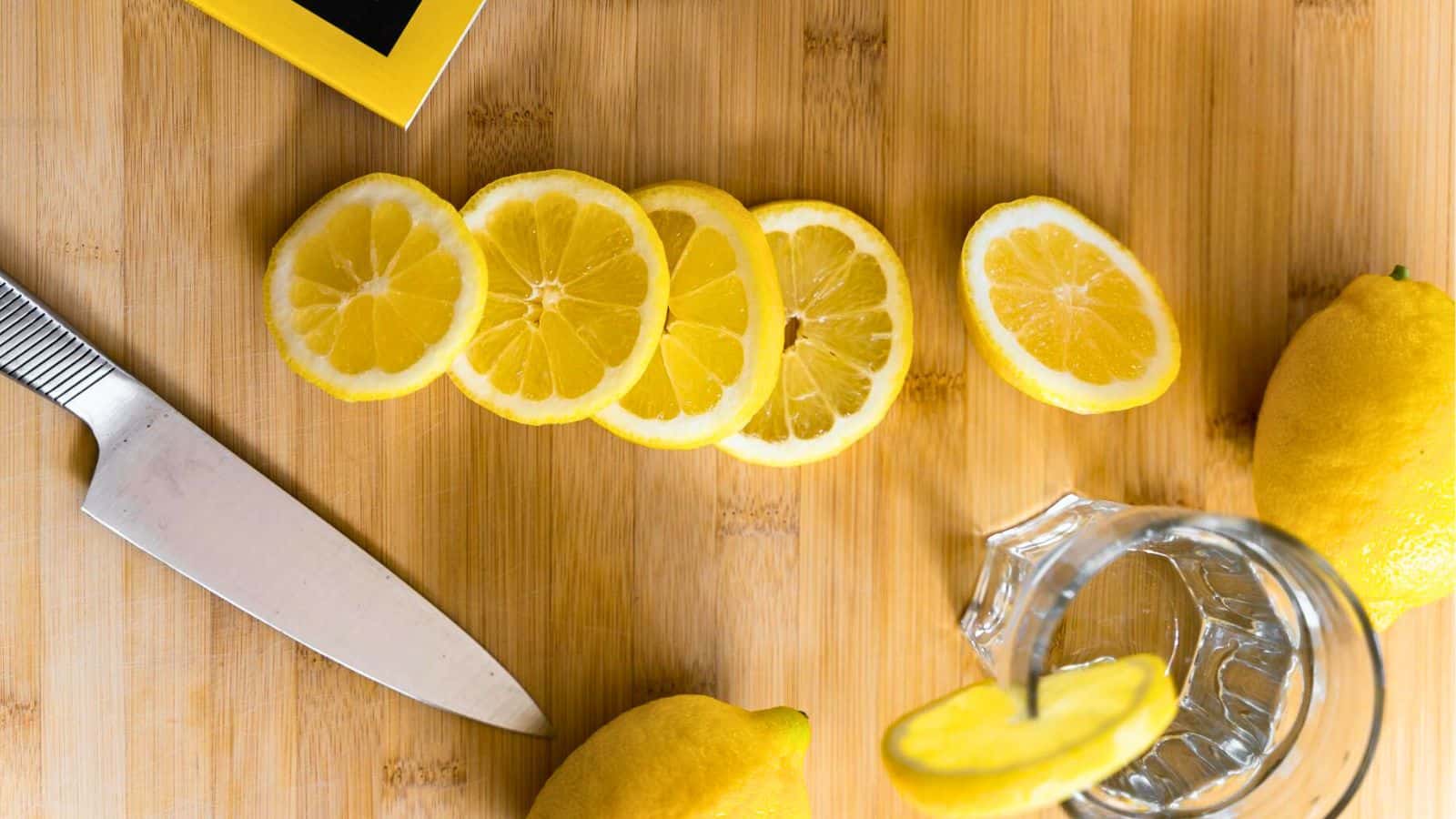 A wooden cutting board with a sliced lemon, a whole lemon, and a knife. A glass with a slice of lemon on its rim is nearby. A yellow-bordered notebook is partially visible in the corner.