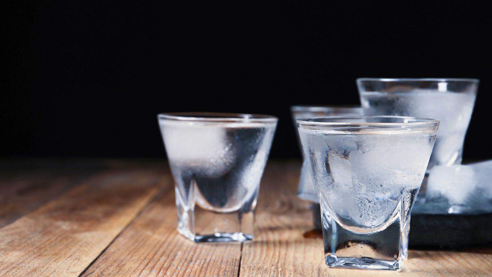 Four frosty shot glasses filled with a clear liquid rest on a wooden table. The background is dark, emphasizing the condensation on the glasses. Two glasses are prominently in the foreground, with two more slightly blurred in the background.