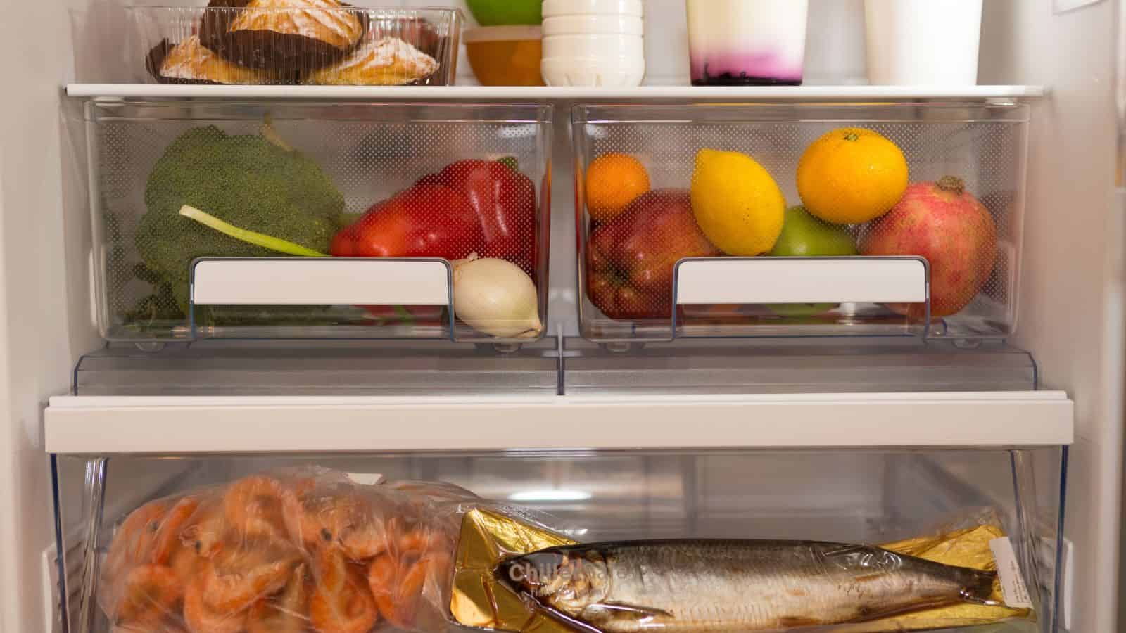 An open refrigerator displaying an assortment of food: the top shelf holds pastries, broccoli, red peppers, a leek, various fruits including oranges and mangoes; the lower shelf contains a bag of shrimp and a wrapped whole fish.