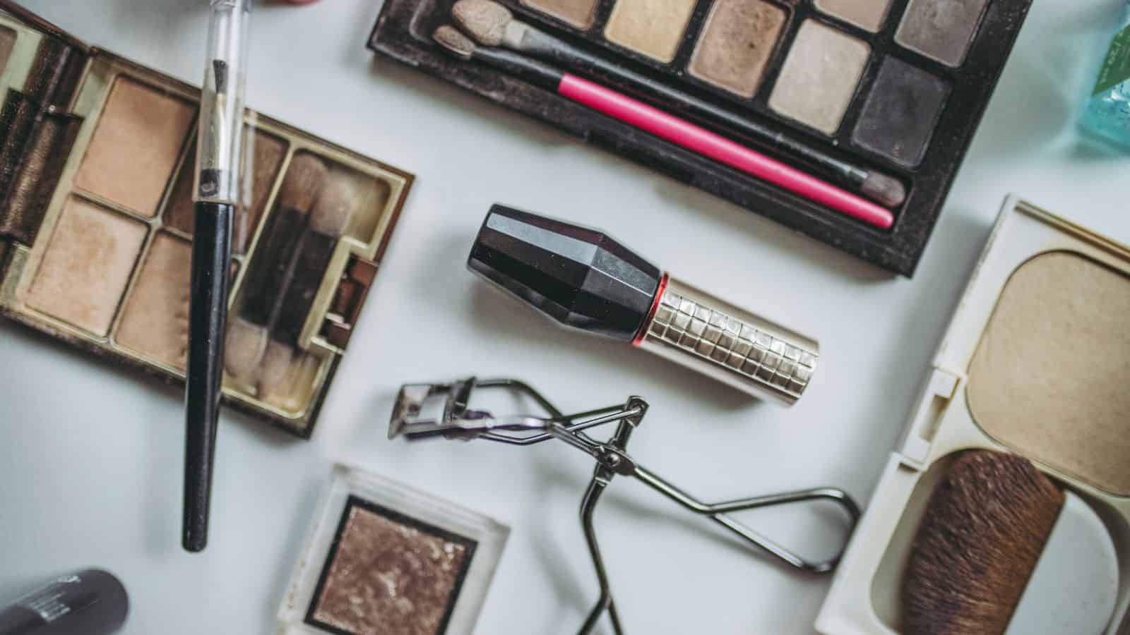 A flat lay of assorted makeup items on a white surface. Includes eyeshadow palettes, a lipstick tube, a mascara wand, an eyelash curler, and brushes. The items are organized in a casual, scattered arrangement.
