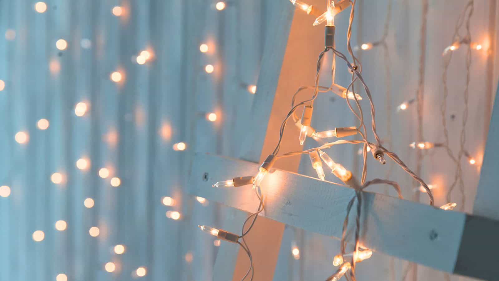 Close-up of warm, glowing string lights against a wooden wall. Some lights are draped over a wooden ladder, creating a cozy and festive ambiance. The background is softly blurred with more lights visible.
