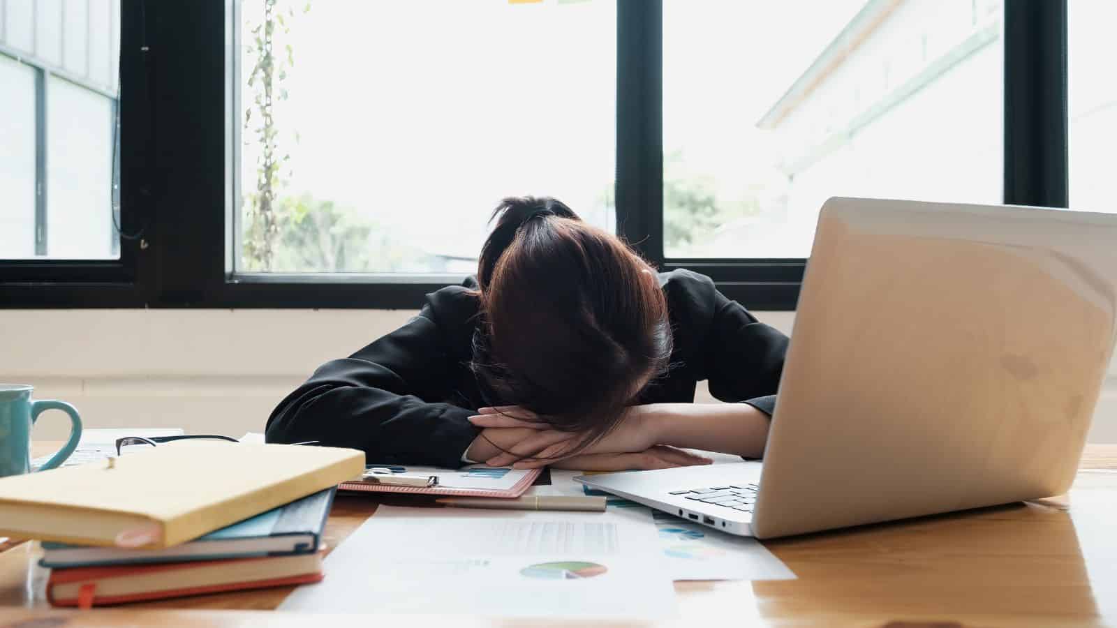 A person with brown hair rests their head on their arms on a desk cluttered with papers, a laptop, books, and a blue coffee mug. Sunlight streams in through large windows in the background.