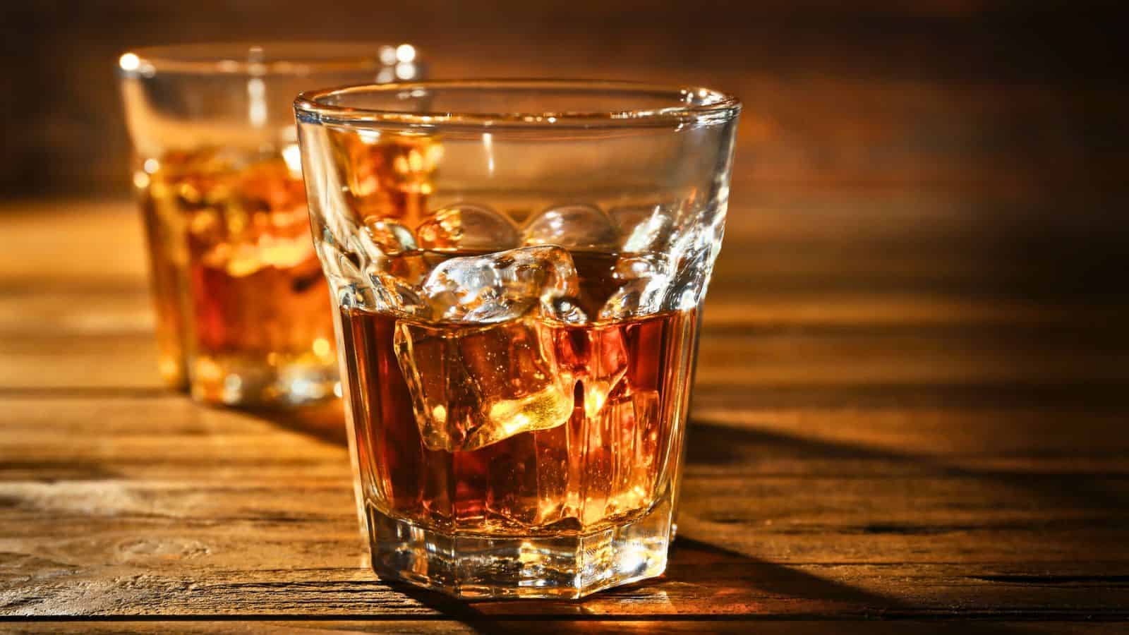 Two glasses filled with amber-colored whiskey and ice cubes sit on a wooden surface. One glass is in the foreground, while the other is slightly blurred in the background. Warm lighting highlights the texture of the liquid and the wood grain.