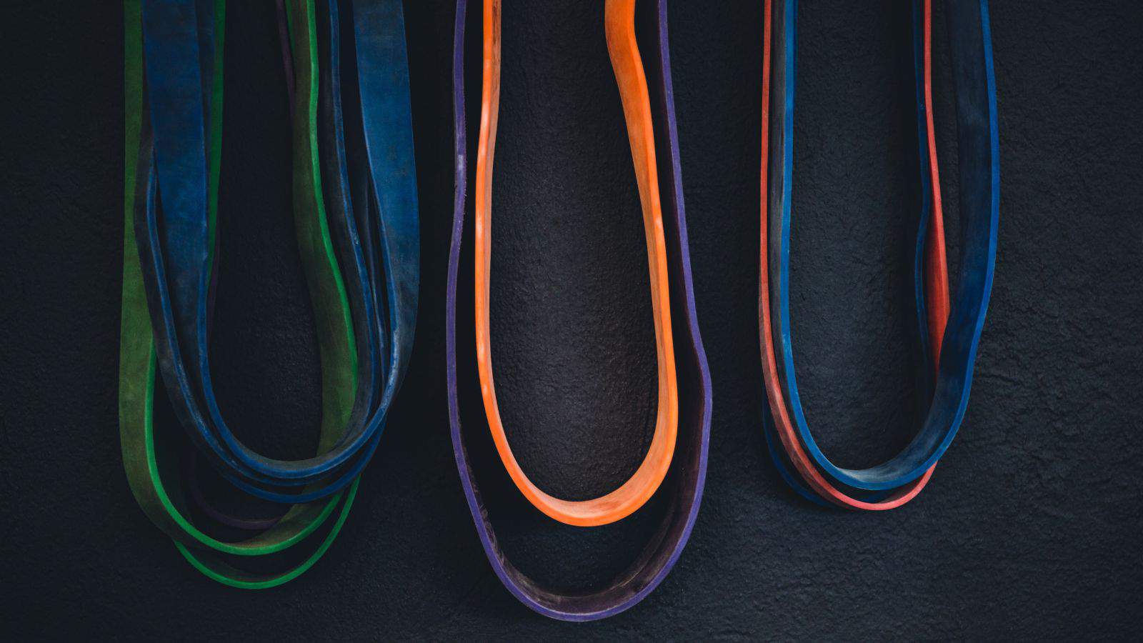 Colorful resistance bands hanging against a dark background. The bands are blue, green, and orange, each looped and stretched vertically.