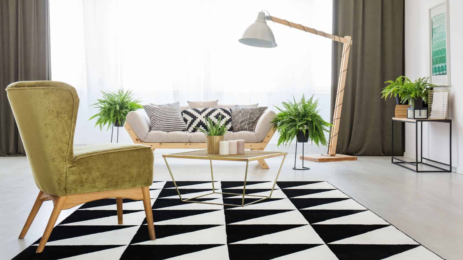 A modern living room with a green armchair, a sofa with patterned cushions, a geometric black and white rug, and a large angled floor lamp. Green plants are placed around the room, and large windows provide natural light.