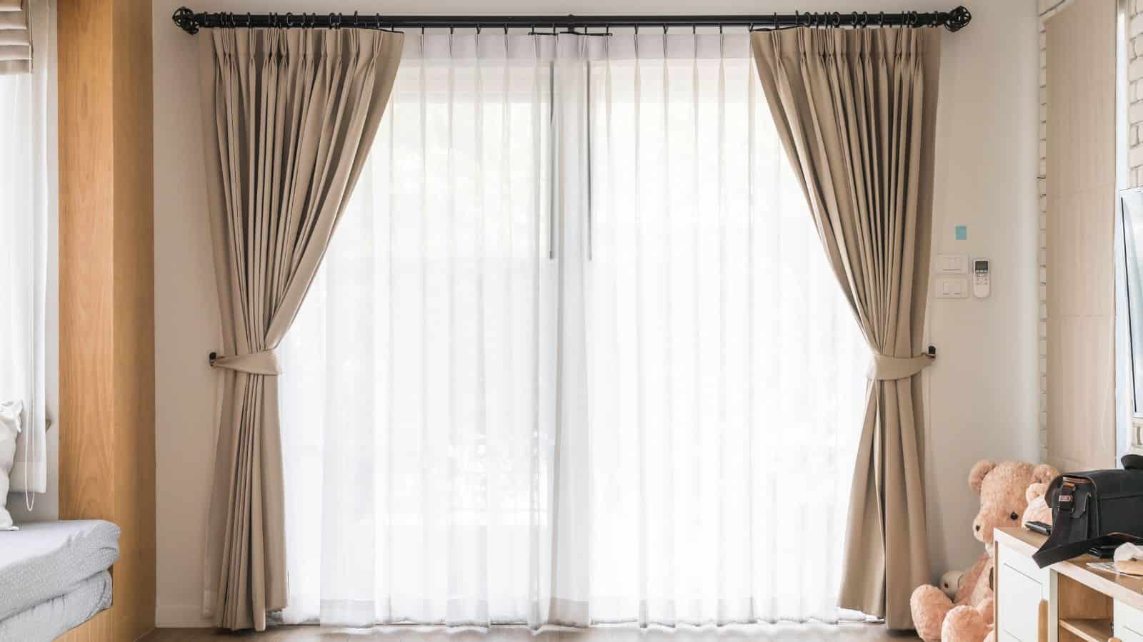 A living room with sheer white curtains lined with beige drapes hanging on a black curtain rod. Two plush bears sit on the right near a white table with various items. Light filters through the window, illuminating the space.