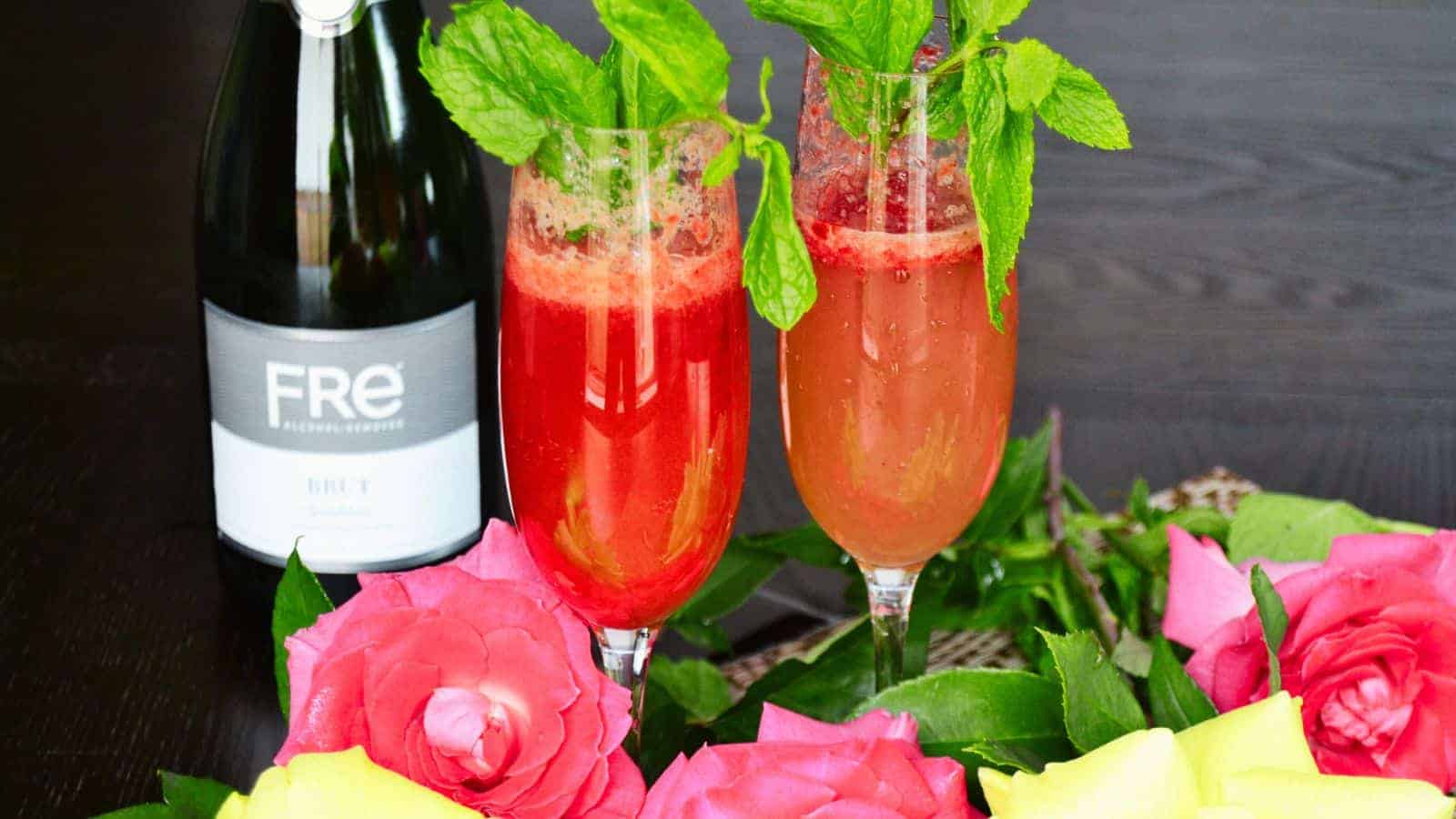 Two red cocktails garnished with mint in champagne glasses are displayed next to a bottle labeled "FRE." The arrangement is surrounded by vibrant pink and yellow roses on a dark surface.
