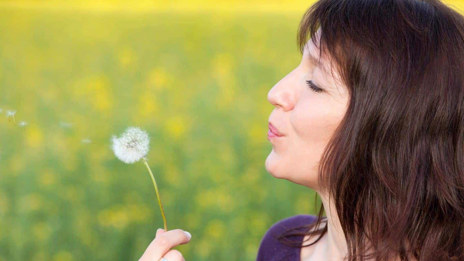 A woman with brown hair is blowing on a dandelion seed head. She stands in profile against a blurred background of green grass and yellow flowers.