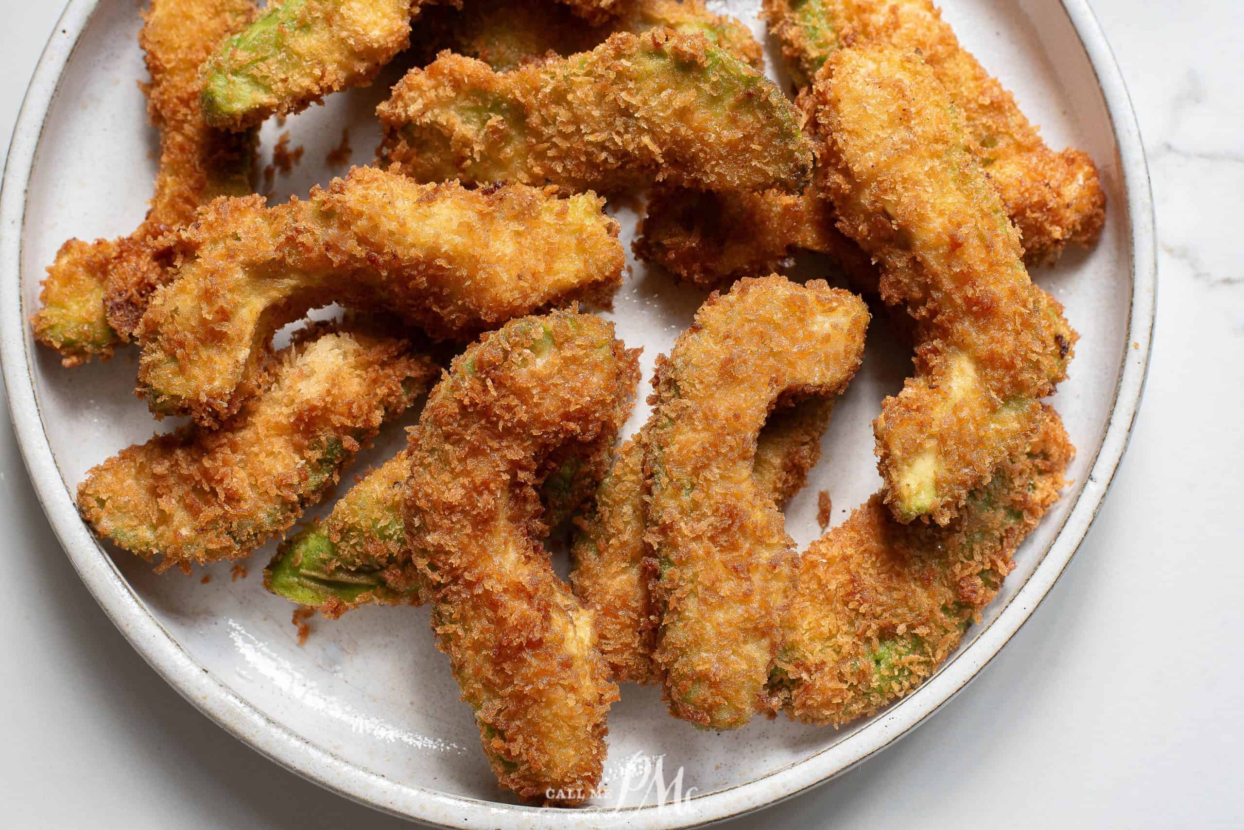 A plate of golden brown, crispy avocado fries is arranged on a white ceramic dish. The fries are breaded and appear freshly fried, showcasing a textured coating. The background is a white marble surface.