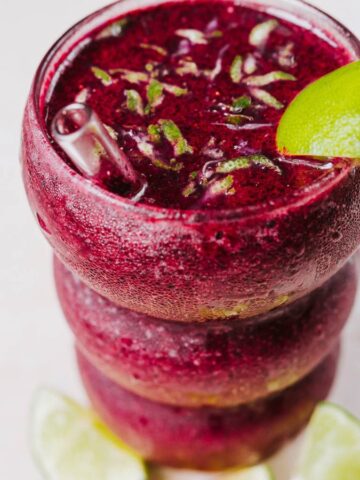 A vibrant purple smoothie in a textured glass, garnished with a lime wedge. The drink has small pieces of herbs on top and is placed on a light surface with two lime slices in the foreground.