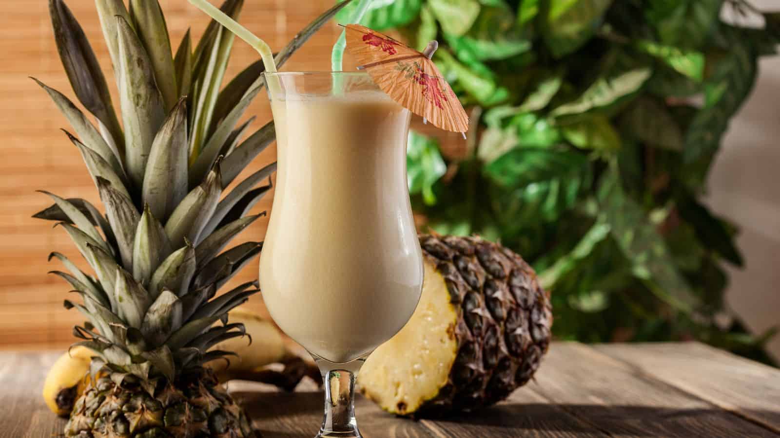 A tall glass of pina colada with a small paper umbrella and two straws, placed on a wooden surface. A whole pineapple and a sliced pineapple are next to the glass. Green leafy background adds a tropical feel.