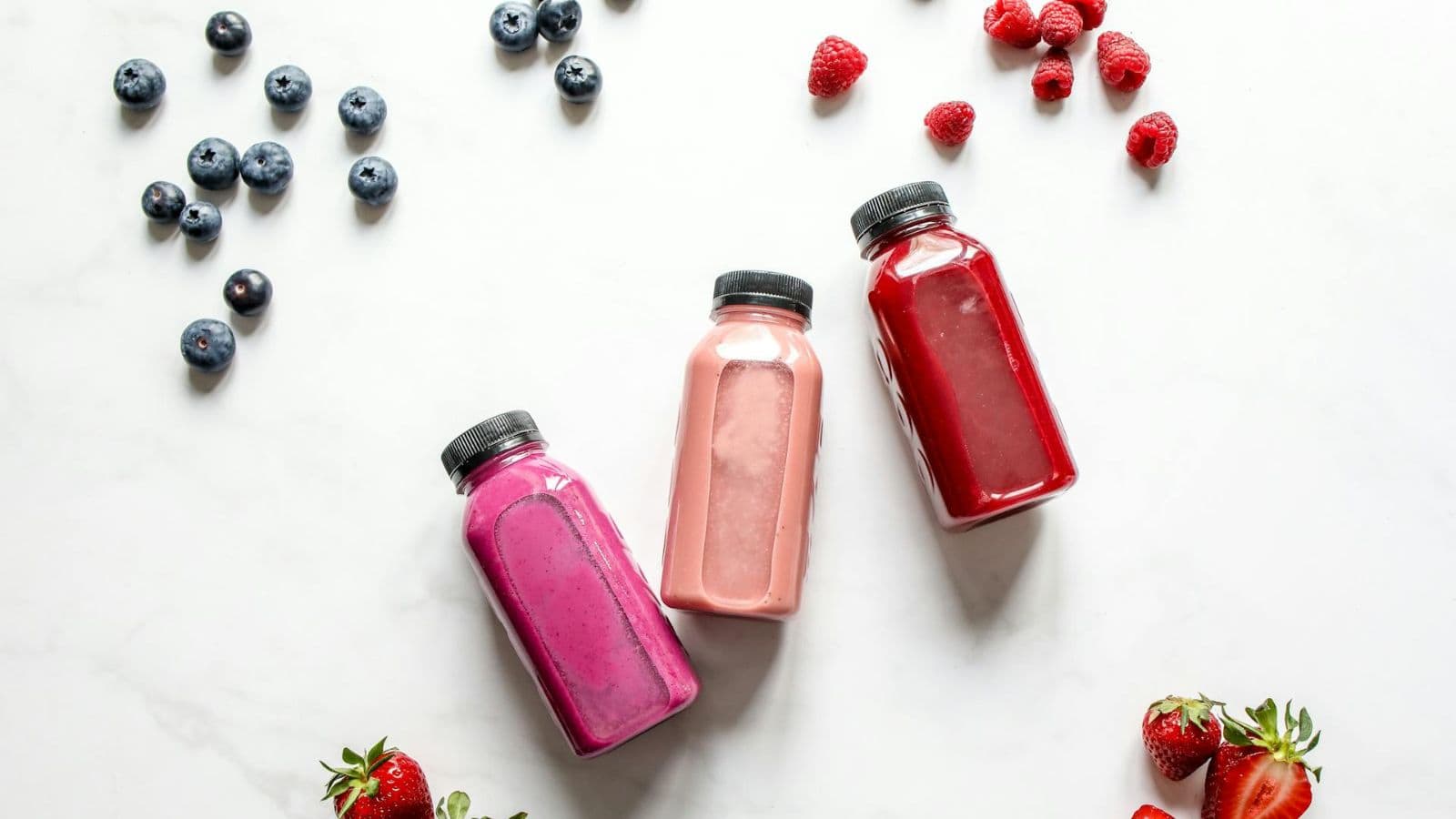 Three bottles of smoothies in purple, pink, and red shades are arranged on a white surface. Surrounding them are scattered blueberries, raspberries, and strawberries.