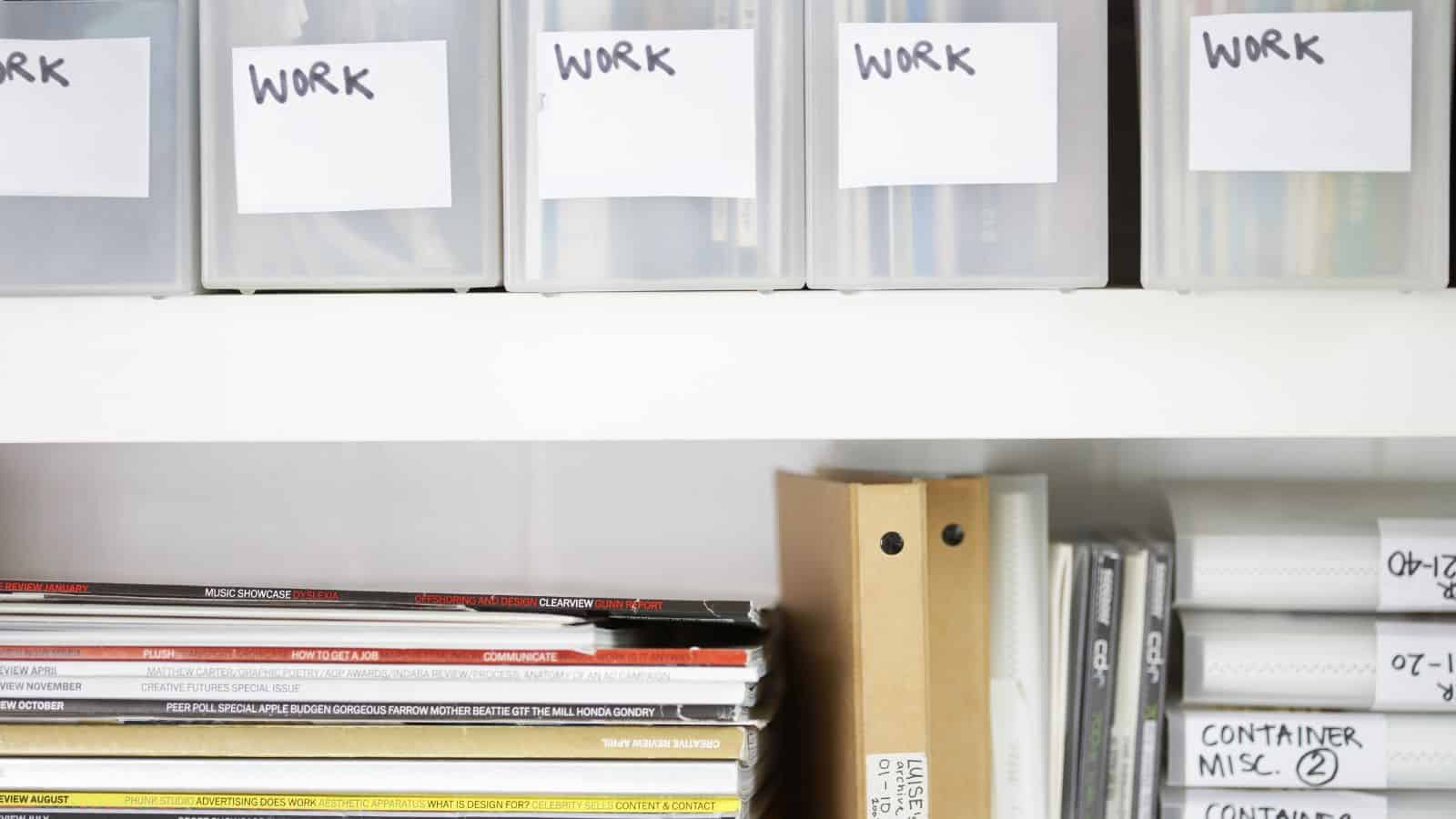 Plastic containers labeled "Work" are on a shelf above neatly stacked magazines and binders. Binders on the right have handwritten labels with dates and descriptions. The scene appears organized and tidy.