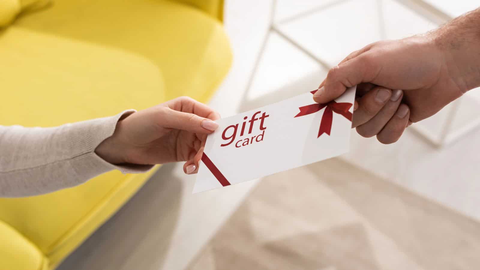 A person hands a gift card in an envelope to another person. The envelope is white with red text and a ribbon design. A yellow cushion and a beige carpet are visible in the background.