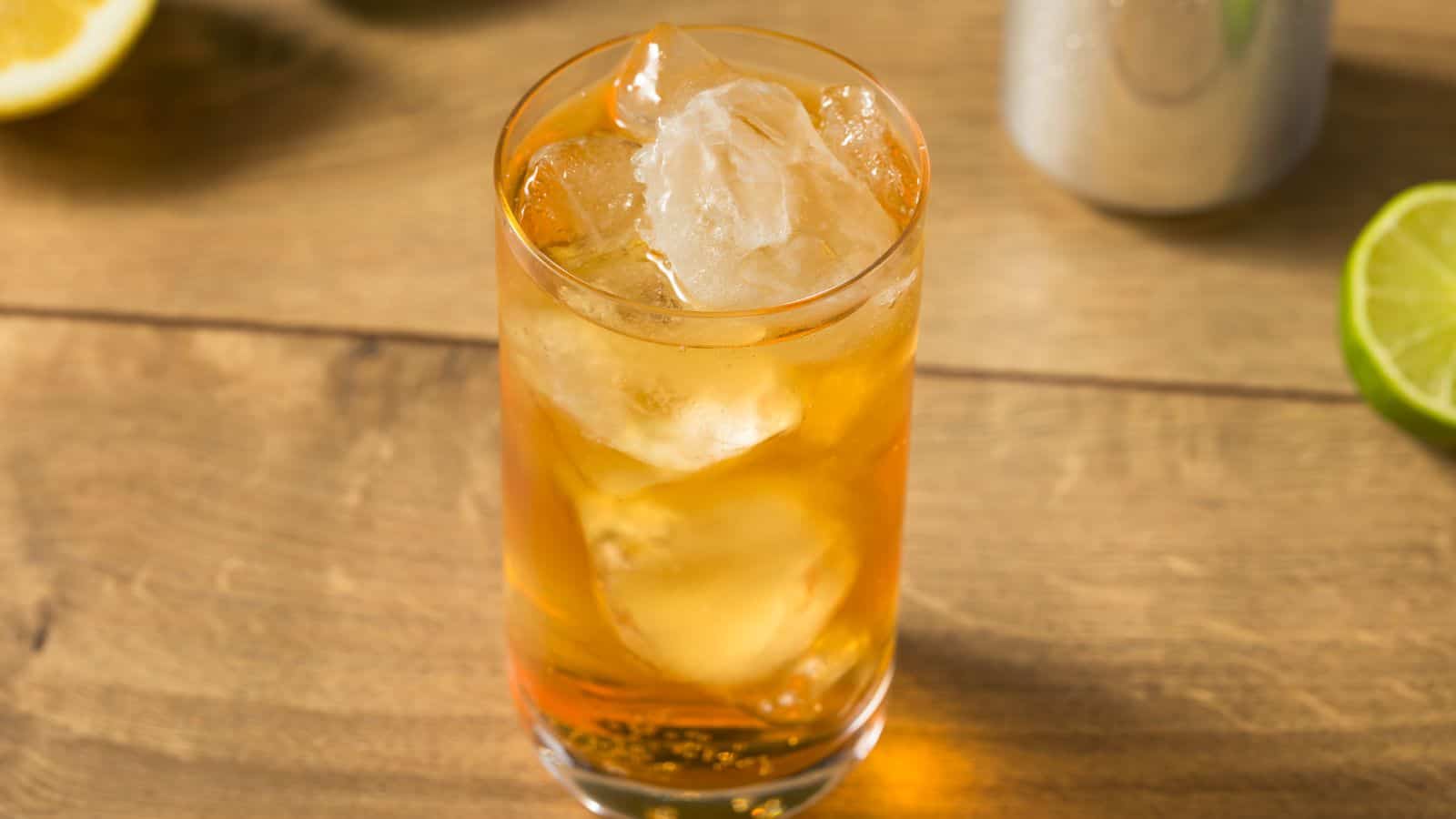 A glass of iced tea sits on a wooden table. The drink is filled with large ice cubes. Partial views of a lemon slice and a lime wedge are in the background.