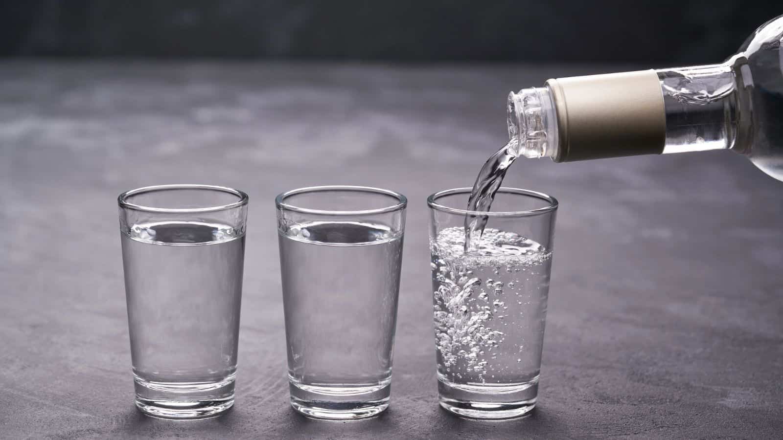 Three clear glasses are aligned on a dark surface. A liquid is being poured into the middle glass from a bottle on the right. The two glasses on the left are filled with a similar clear liquid.