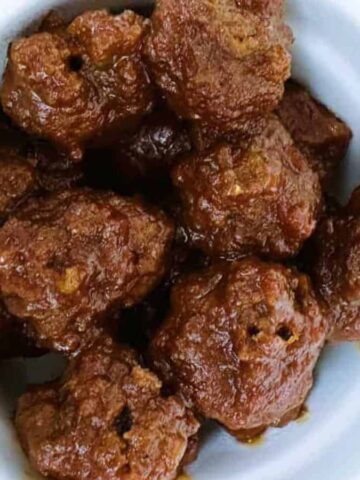 A white bowl filled with cooked meatballs coated in a brown sauce, placed on a wooden surface. A portion of a yellow cloth is visible in the top right corner.