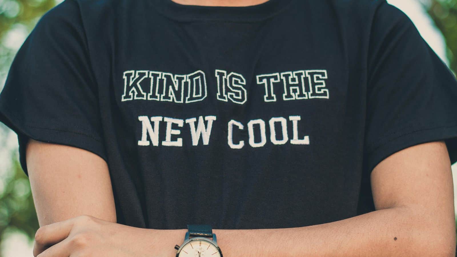 Person wearing a black T-shirt with the text "KIND IS THE NEW COOL" in white letters. Arms are crossed, and a watch is visible on the left wrist. The background is blurred greenery.