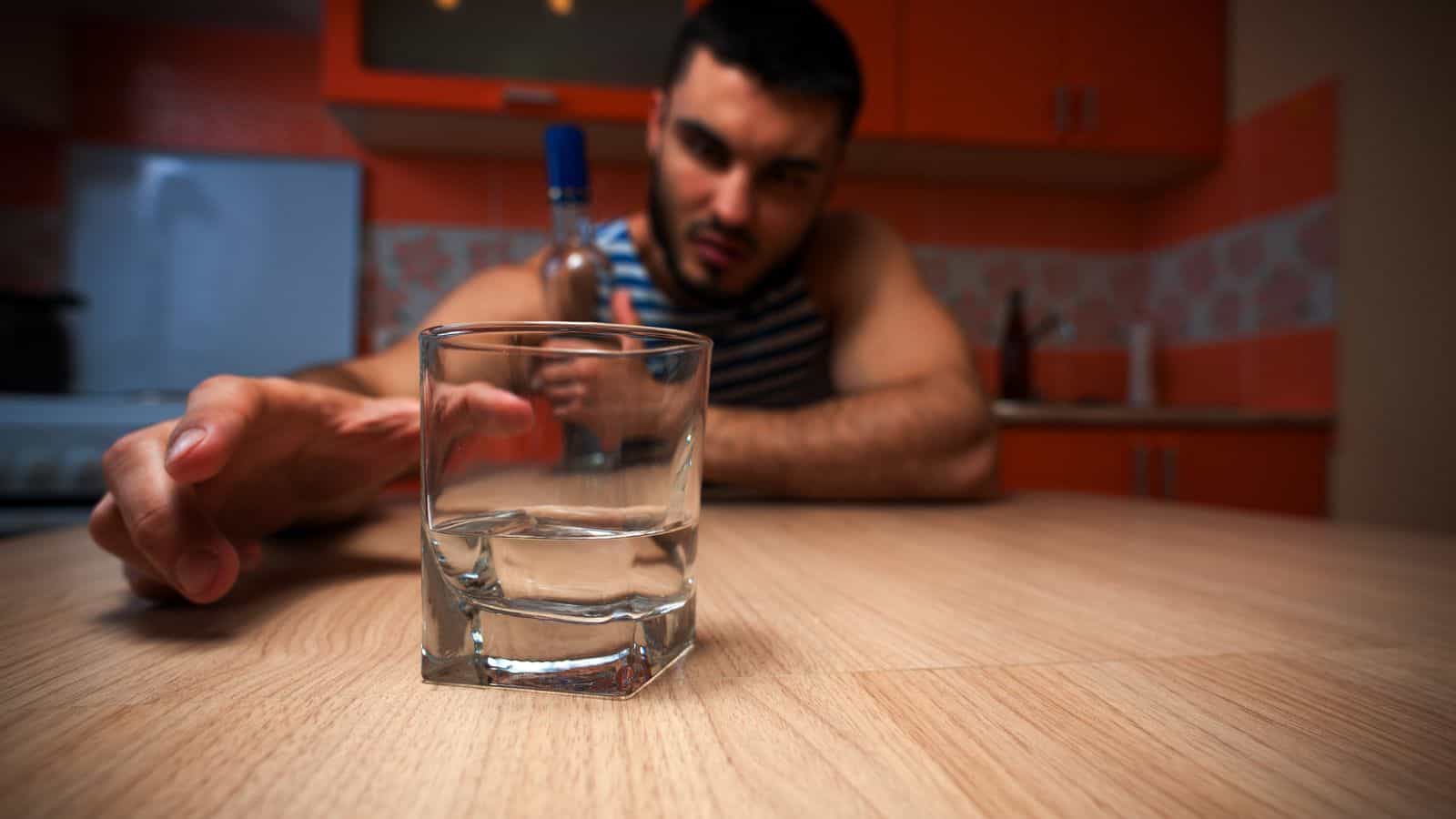 A man sits at a kitchen table with a focused expression, holding a bottle, reaching towards a nearly empty glass in the foreground. The kitchen has orange cabinets and a tiled backsplash.