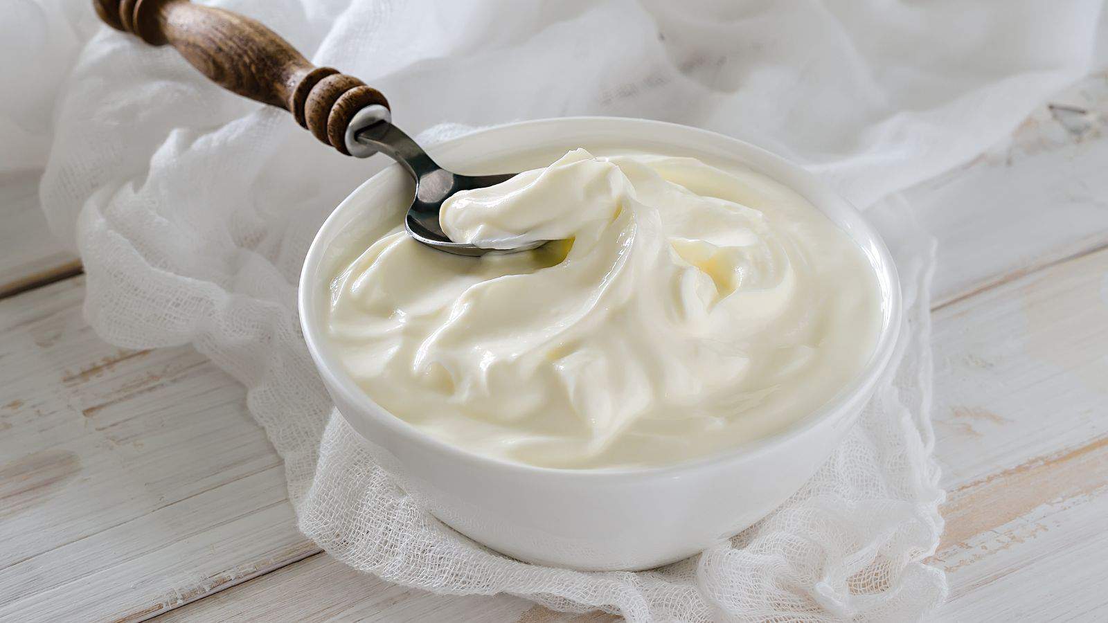 A bowl of creamy white yogurt with a spoon in it, placed on a wooden surface. A white cloth is seen underneath the bowl. The yogurt appears smooth and freshly served.
