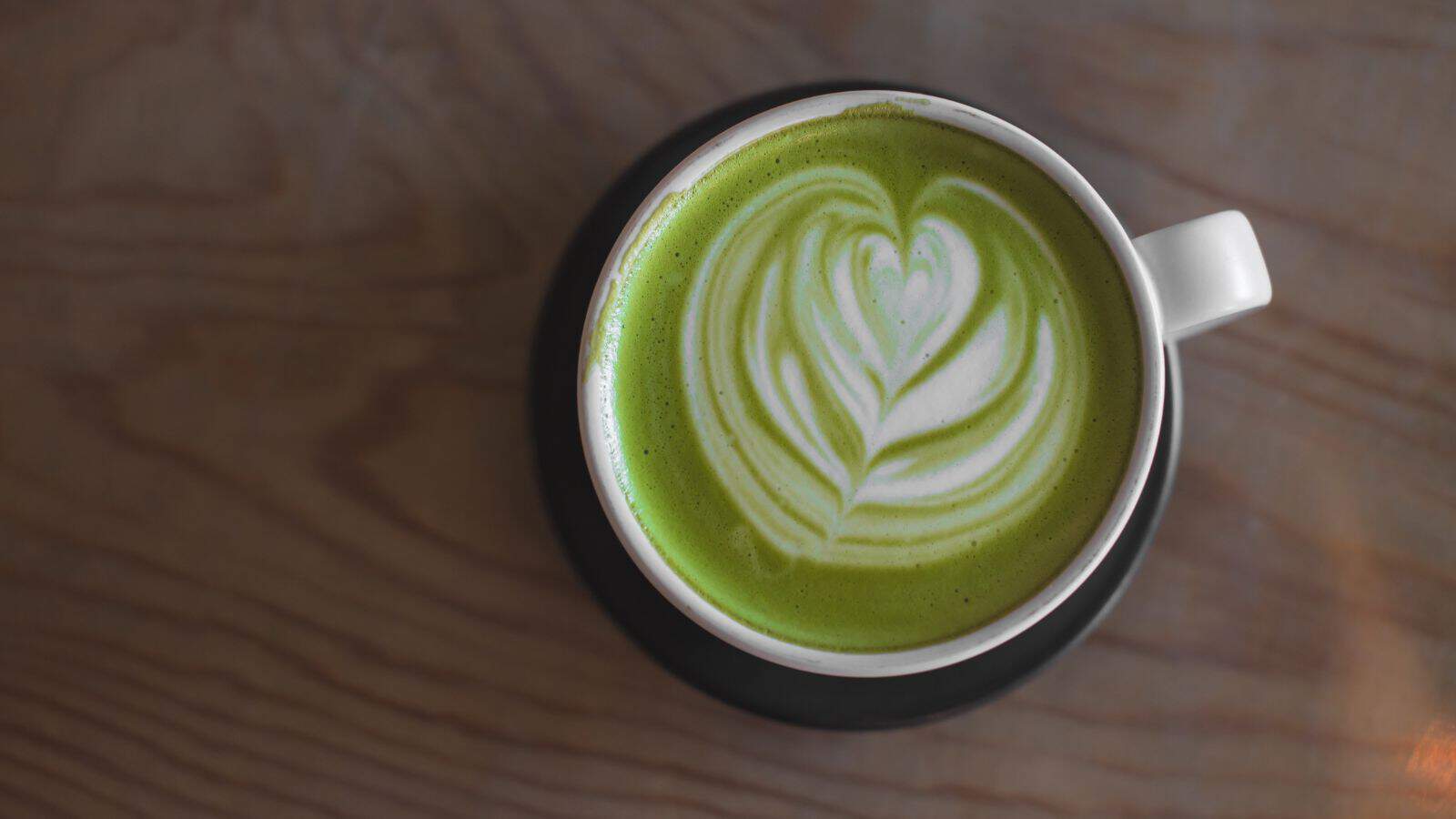 A top view of a cup of matcha latte on a black saucer, placed on a wooden surface. The green beverage is topped with intricate white latte art resembling a heart shape.