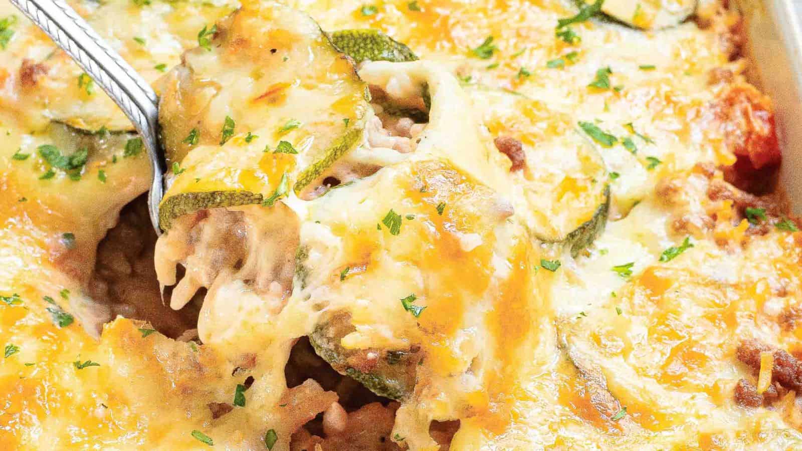 Close-up of a cheesy zucchini casserole being served with a spoon. The dish features layers of melted cheese, sliced zucchini, ground meat, and herbs, with a golden-brown crust.