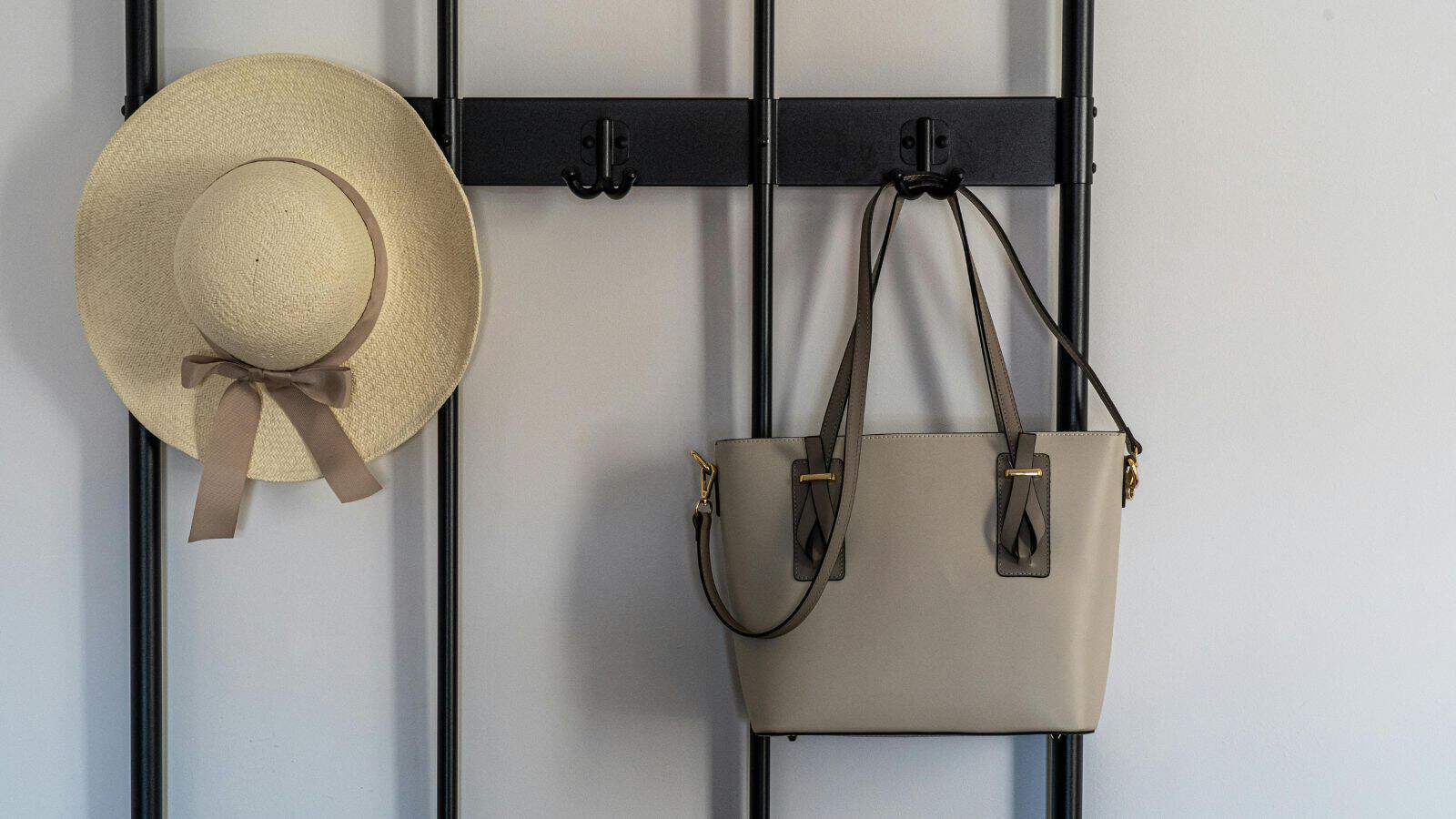 A beige straw hat with a bow and a taupe handbag hang on a black metal wall rack against a light gray background.