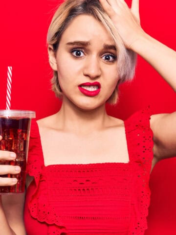 A person with short hair and a worried expression holds a plastic cup with a straw containing a dark liquid. They wear a red top and stand against a red background, with one hand on their forehead.