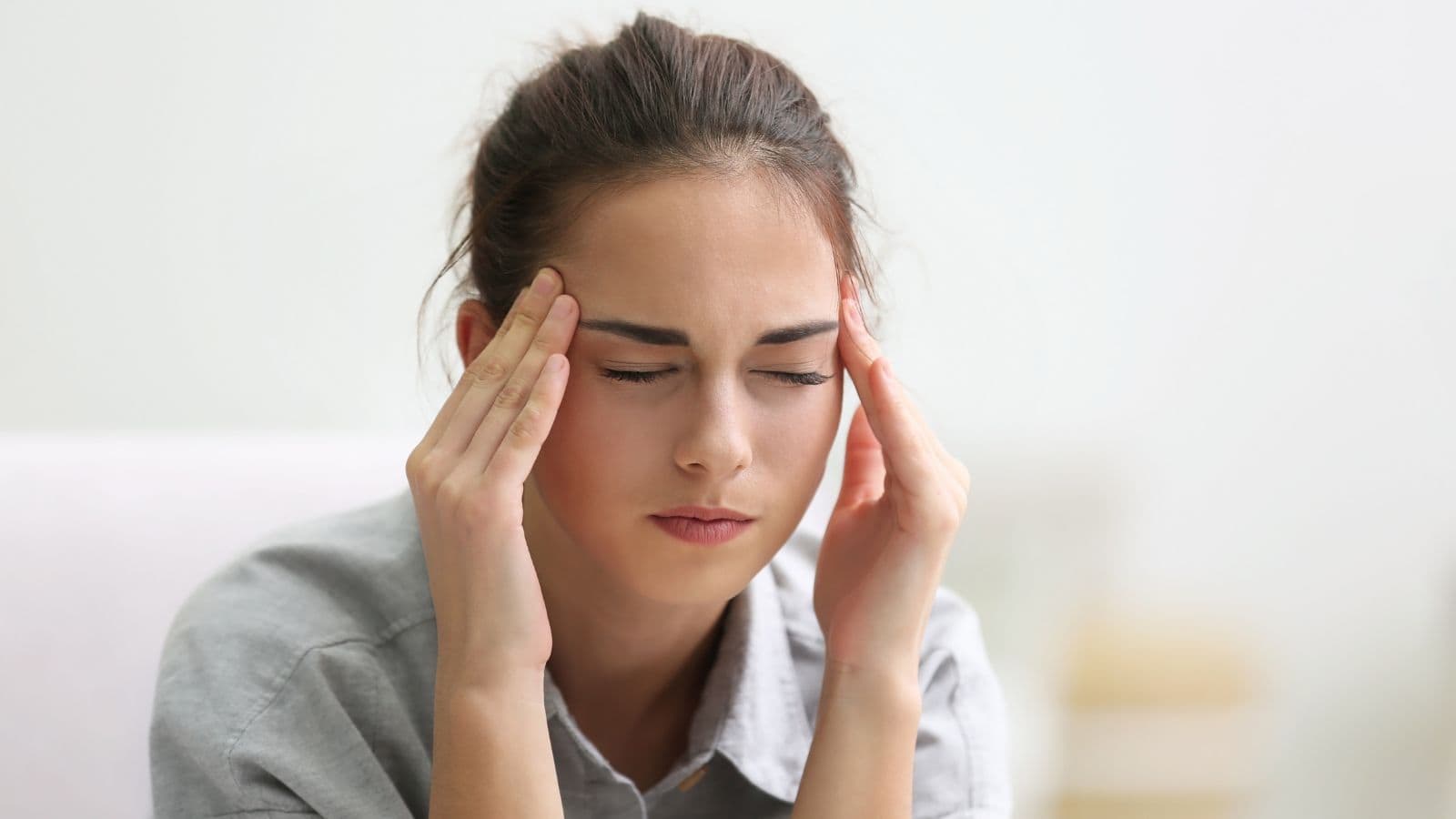A woman with closed eyes is pressing her temples with her fingers, appearing to be in discomfort. She is wearing a gray shirt and has dark hair tied back. The background is softly blurred.