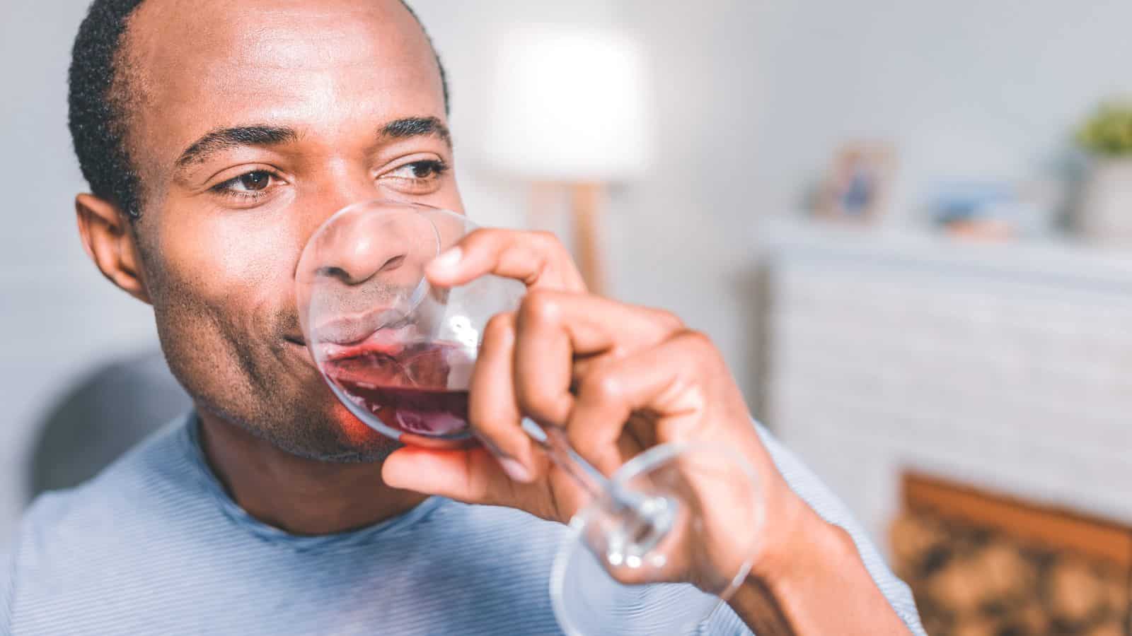 A person drinks a glass of red wine while sitting in a room with a cozy atmosphere. There is a lit lamp and some decorations visible in the background. The person appears relaxed and contemplative.